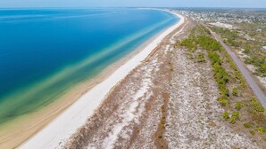 An aerial view of a coastal area with a long sandy beach, a road parallel to the shore, and a distant town with buildings. - Sealavie (Port St. Joe)