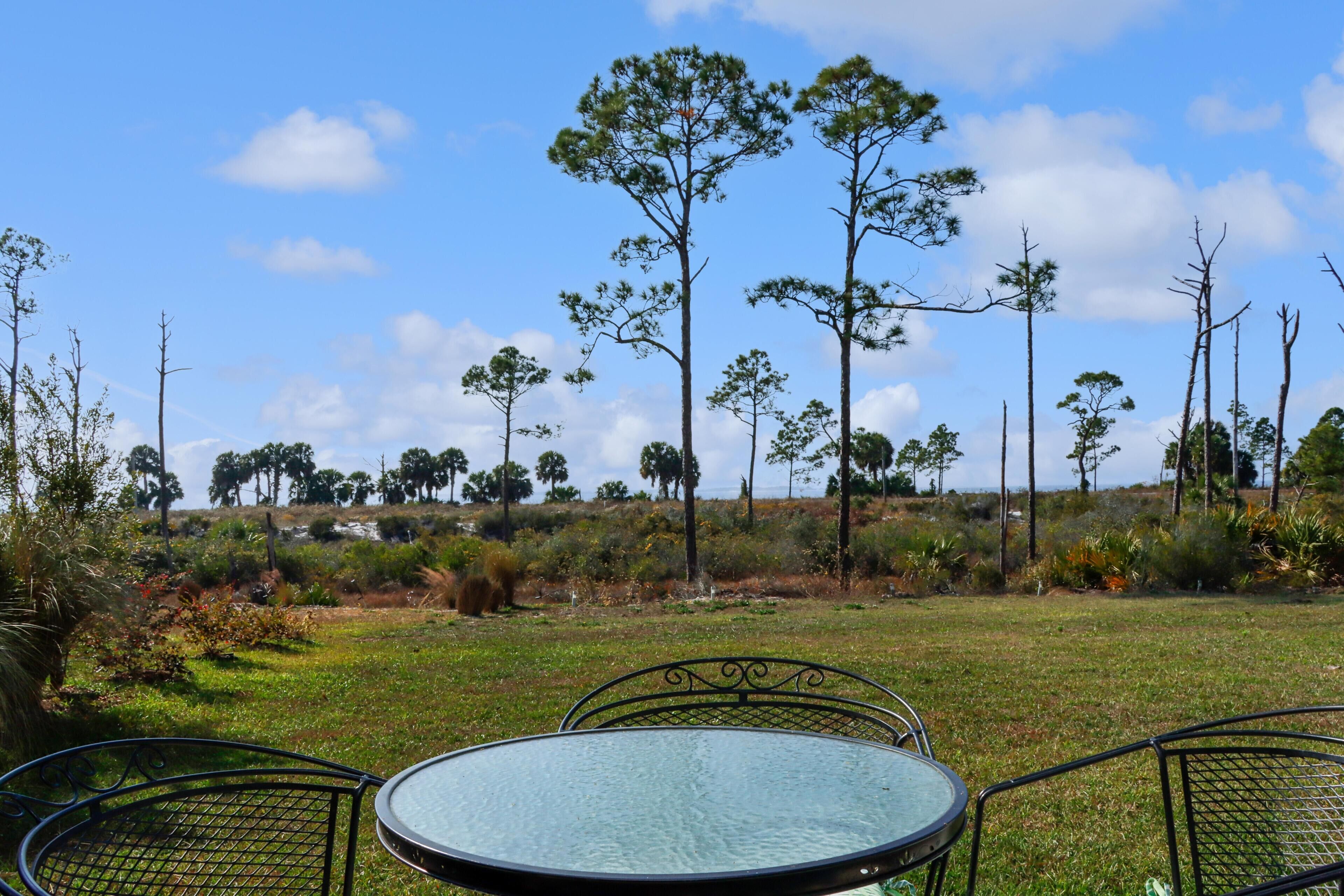 A round glass table with metal chairs in a grassy outdoor area with trees in the background.