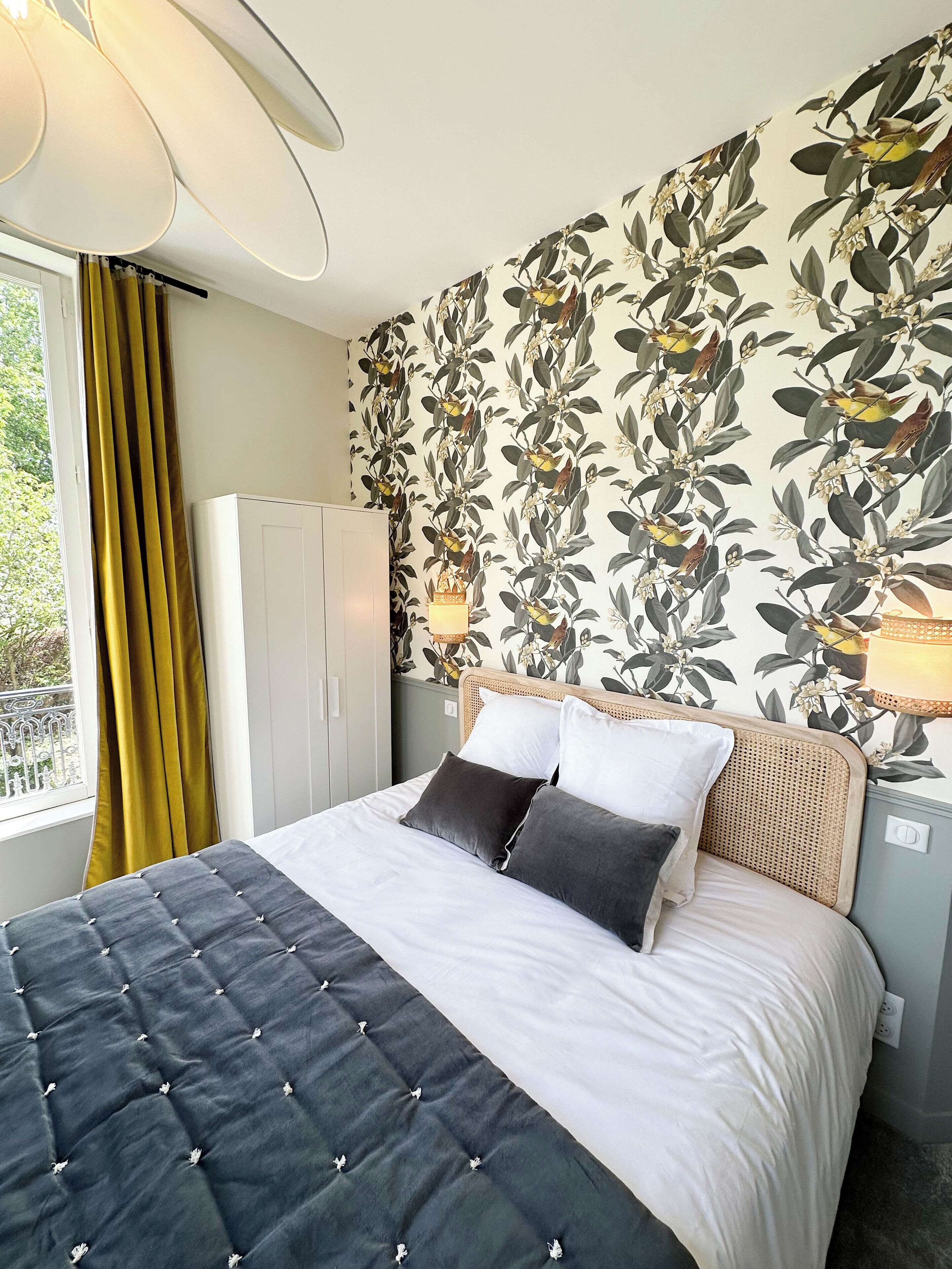 A bedroom with a bed, a wardrobe, a window with curtains, and a wall with a floral wallpaper.