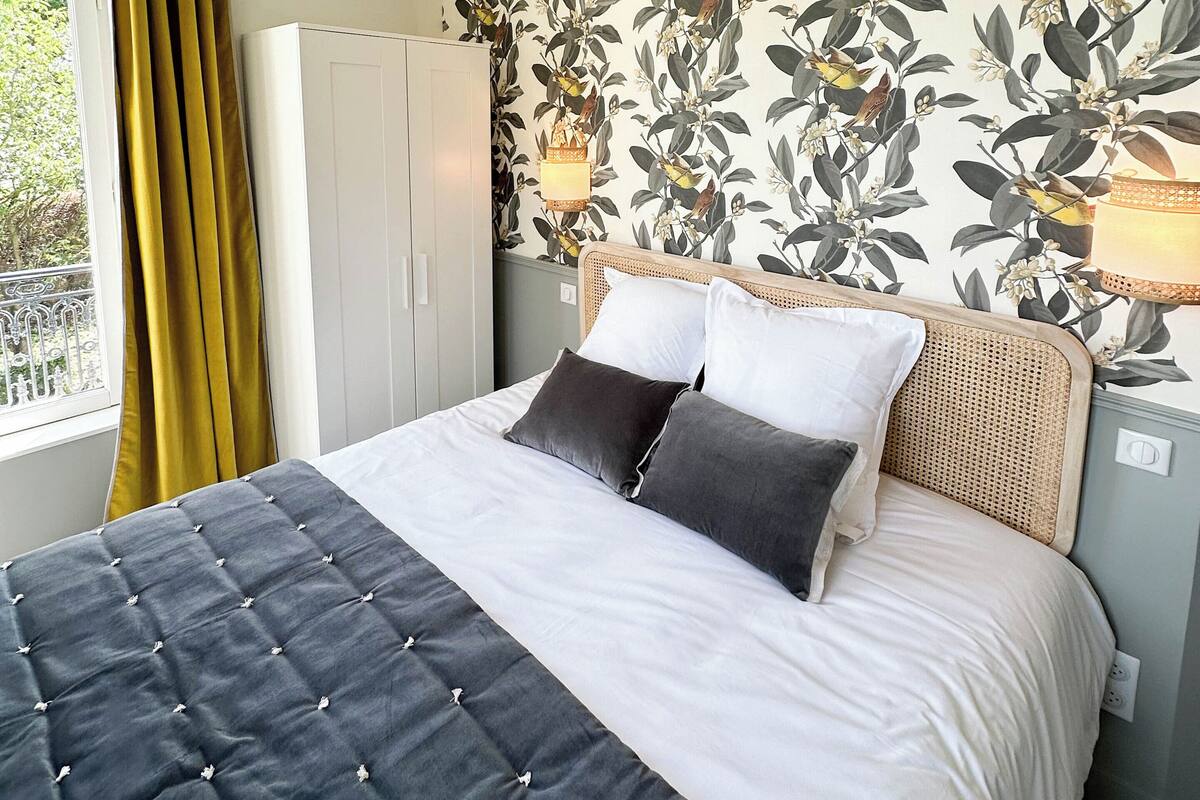 A bedroom with a bed, a wardrobe, a window with curtains, and a wall with a floral wallpaper.