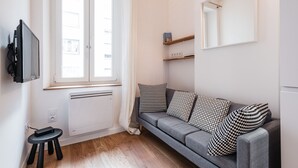 A modern living room with a grey sofa, black and white patterned cushions, a small black stool, a mounted TV, and a window with white curtains. - SmartStay Perrache - Presqu'île (Lyon)