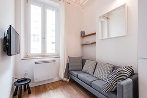 Ein modernes Wohnzimmer mit grauem Sofa, Kissen in Schwarzweißmuster, einem kleinen schwarzen Hocker, einem an der Wand montierten Fernseher und einem Fenster mit weißen Vorhängen.