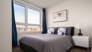 Un dormitorio con cama, mesita de noche, lámpara, ventana con cortinas e imagen colgada en la pared.