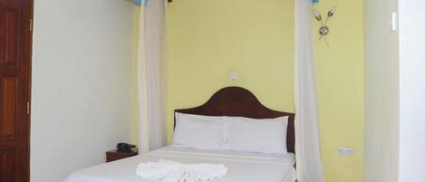Un lit simple avec du linge de lit blanc et une serviette blanche, une tête de lit en bois et un mur jaune avec une fenêtre et un luminaire fixé au mur.