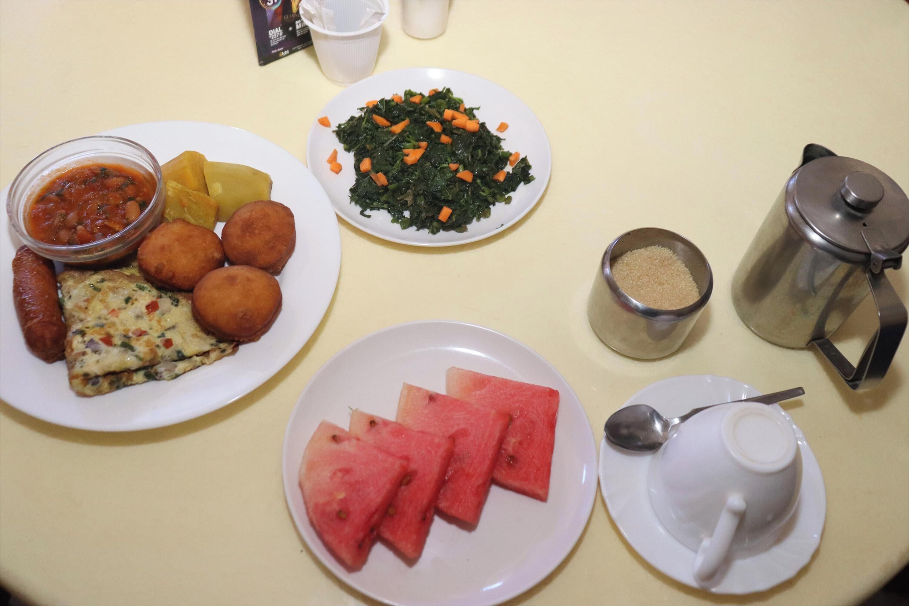 Una mesa con platos de comida, incluyendo una porción de pizza, sandía y un guiso de verduras, además de una cafetera y una taza.