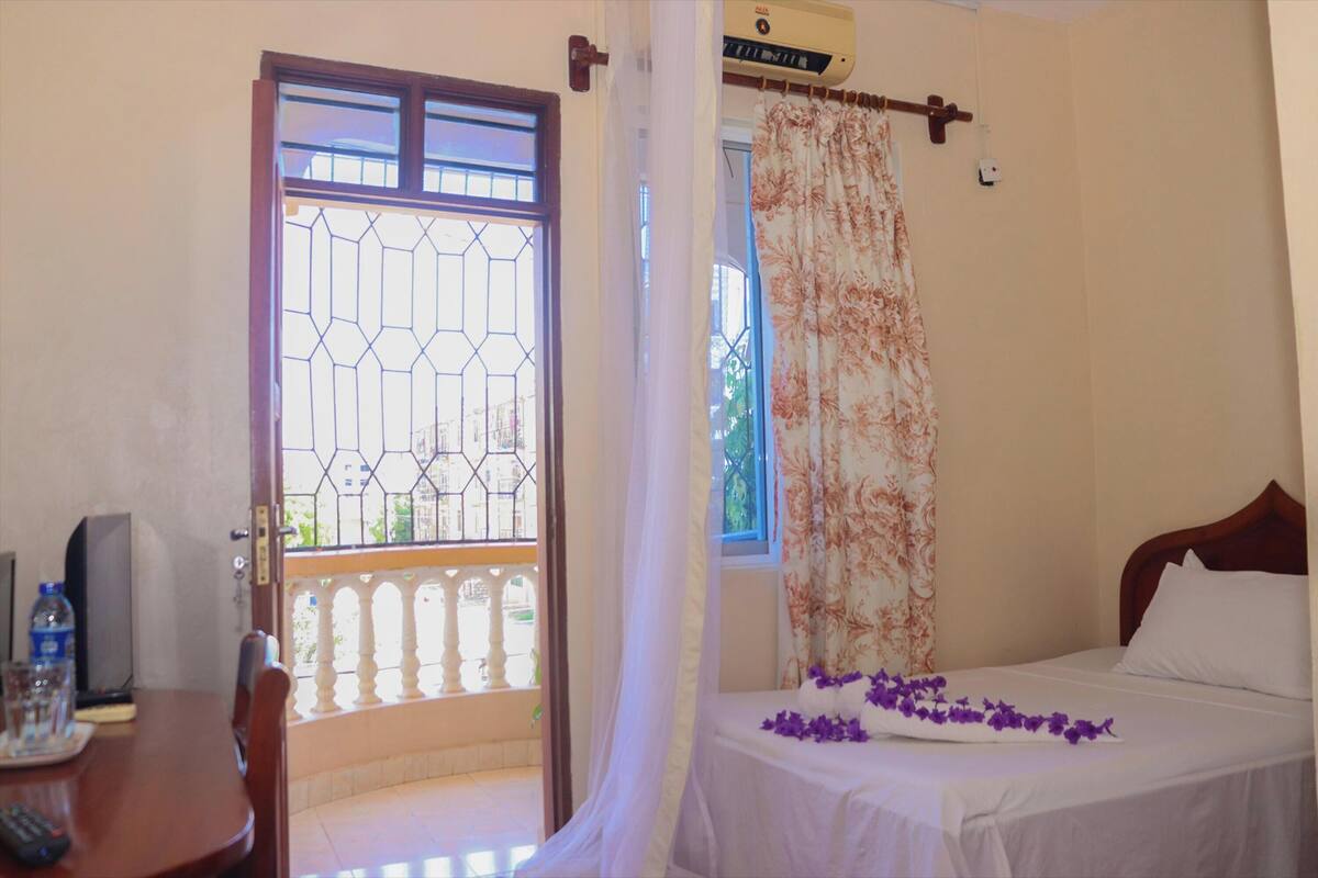A room with a bed, a table with a bottle and glasses, a curtain, and a window with a view.