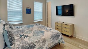 Une chambre à coucher comprenant un lit, deux fenêtres avec stores, une commode, un téléviseur et un ventilateur de plafond.