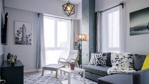 Ett modernt vardagsrum med en grå soffa, ett vitt soffbord, en trästol och en golvlampa. Det finns ett svart TV-bord och en inramad tavla på väggen.