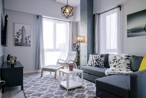 A modern living room with a grey sofa, a white coffee table, a wooden chair, and a floor lamp. There is a black TV stand and a framed picture on the wall. - Buchanan *201* - Midtown Condo Luxe + Gym (Montreal)
