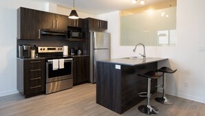 A modern kitchen with dark wood cabinets, stainless steel appliances, a central island with bar stools, and a large mirror above the sink. - Buchanan *609* - Midtown Luxe Loft + gym (Montreal)