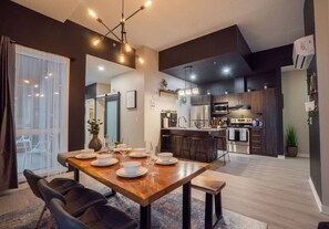 A modern dining area with a wooden table set for four, a kitchen in the background, and a large window with sheer curtains. - Buchanan *304* - Midtown Luxe Condo + gym (Montreal)