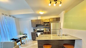 A modern kitchen with a central island, bar stools, a dining area with a table and chairs, and a view of the city through large windows. - Buchanan *705* - Midtown Luxe Loft + gym (Montréal)