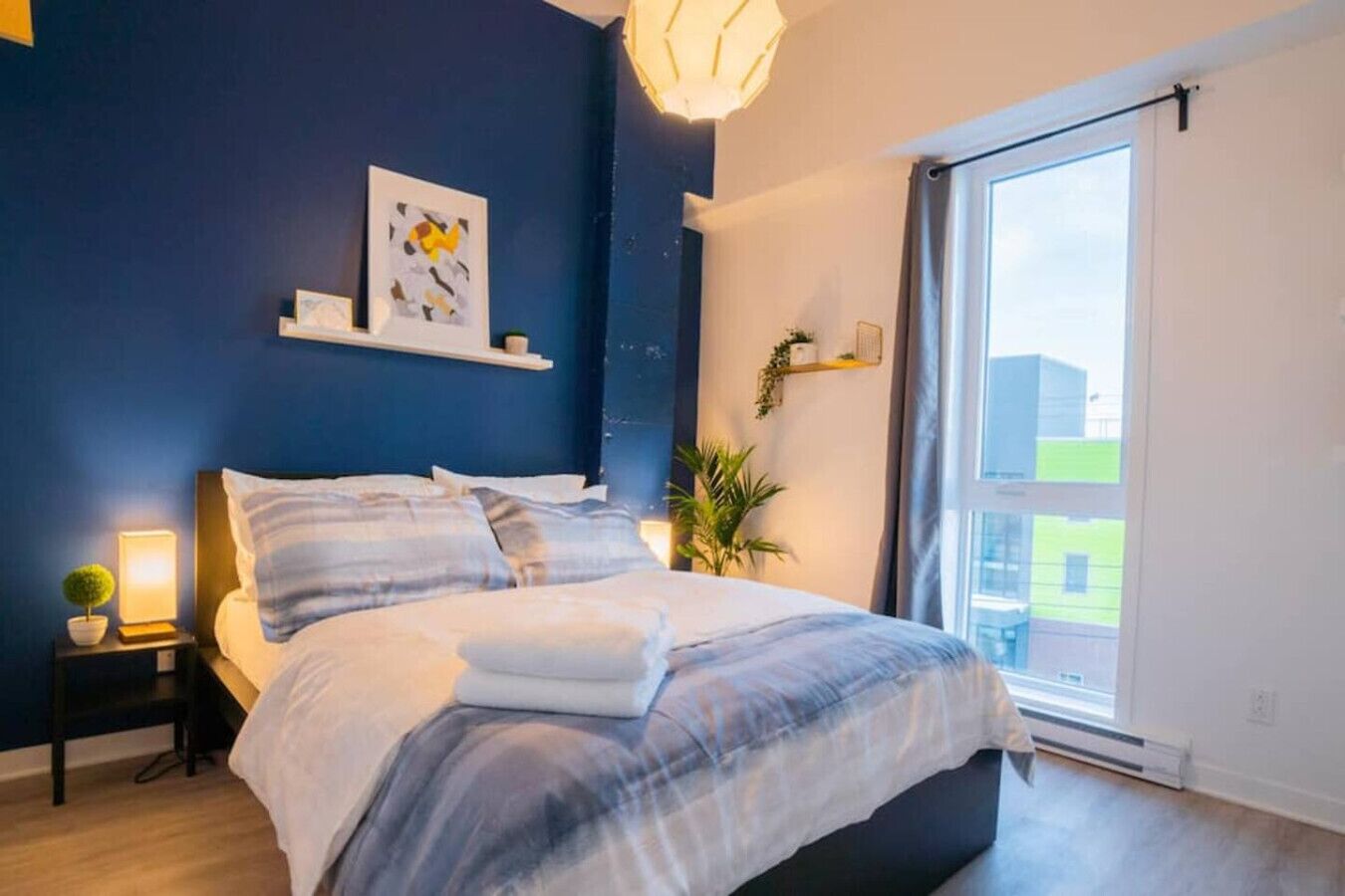 A bedroom with a bed, bedside tables, a nightstand, a window, and a view of buildings outside.