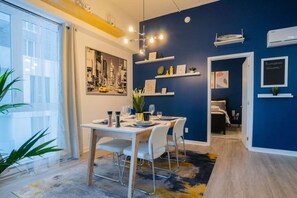 A modern dining area with a table set for two, a cityscape painting, and a blue accent wall. - Buchanan *301* - Midtown Condo Luxe + Gym (Montreal)