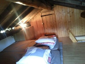 Une petite chambre mansardée, peu éclairée, avec des murs en bois et un plafond en pente. On y trouve un lit avec des oreillers et une couverture, ainsi qu’un banc en bois adossé au mur.