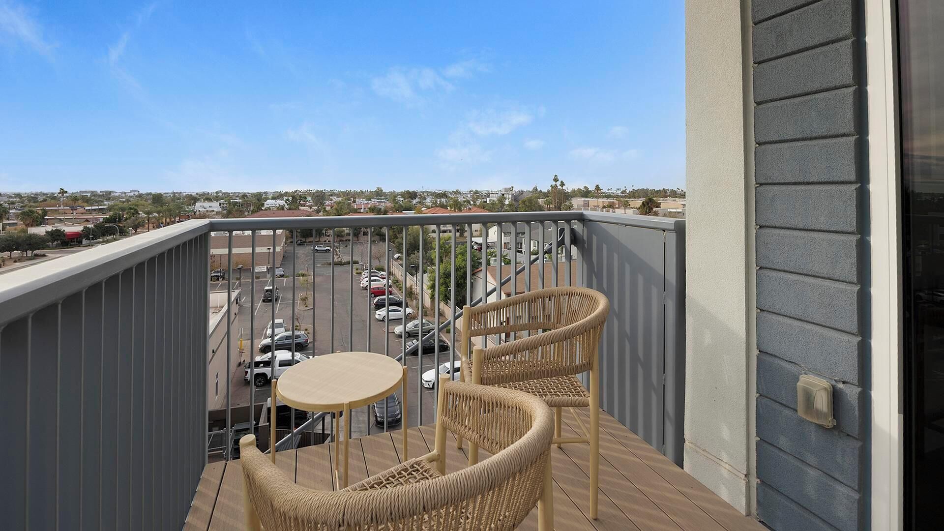 A balcony with wicker chairs and a small table, overlooking a parking lot and buildings.