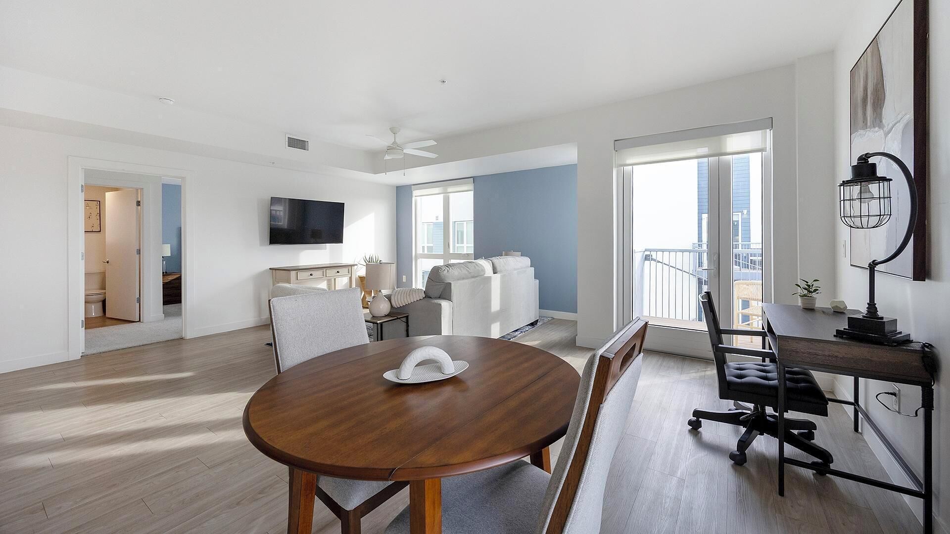 A modern living room with a wooden dining table, a sofa, a TV mounted on the wall, and a balcony with a railing.