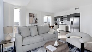 Een moderne woonkamer met een grijze bank, een salontafel en een fauteuil. Er is een keuken met een koelkast, magnetron en spoelbak.