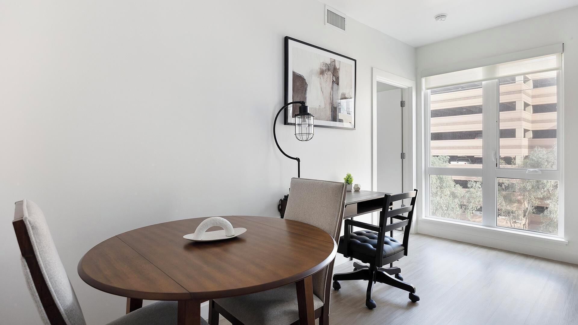 Un comedor moderno con una mesa redonda de madera, dos sillas, una lámpara colgante y vistas a un aparcamiento a través de amplias ventanas.