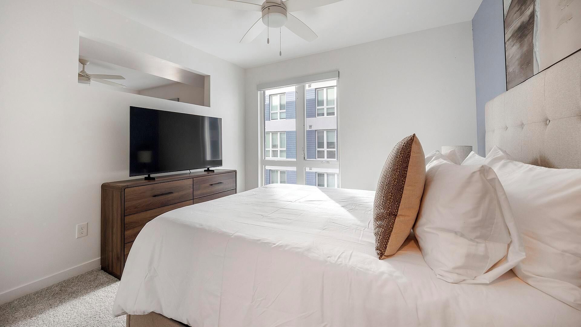 A bedroom with a bed, a television on a wooden dresser, a ceiling fan, and a window with a view of buildings.