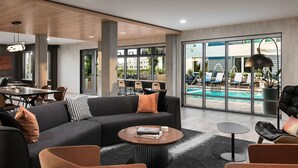 A modern lounge area with a black sofa, orange cushions, a round coffee table, and a pool view through large windows. - Landing | Beautiful Studio, Clubhouse, Yoga Studio (Tempe)