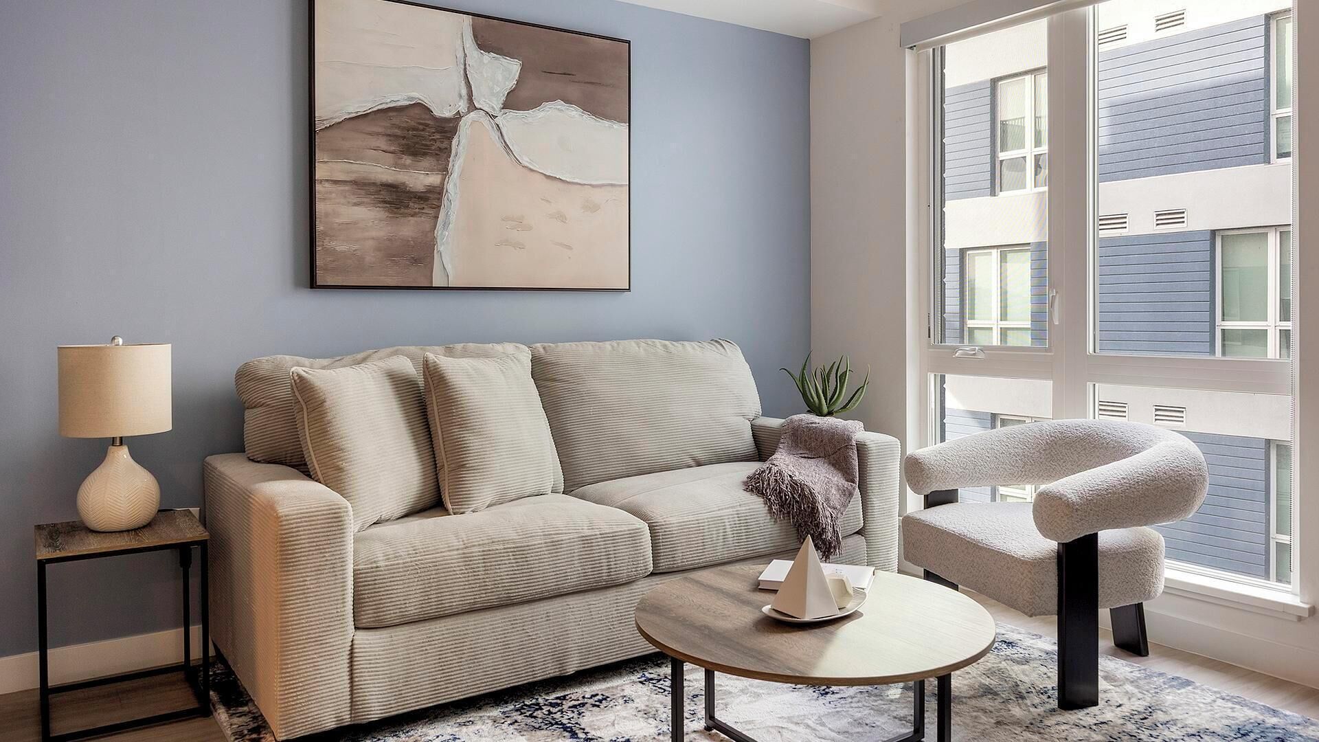 A living room with a beige sofa, a round coffee table, a side table with a lamp, a modern chair, and a large abstract painting on the wall.