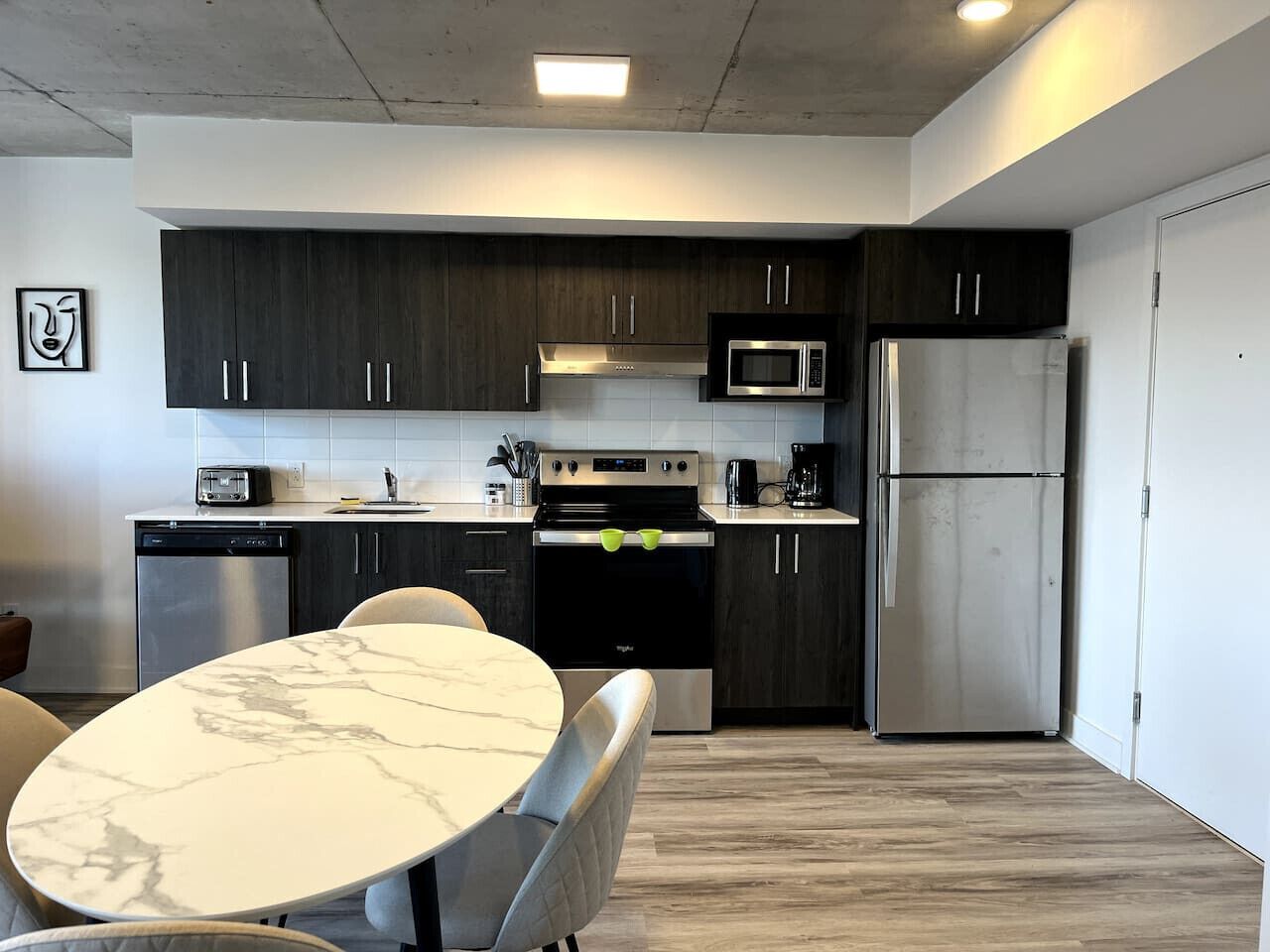 A modern kitchen with dark cabinets, a stainless steel refrigerator, a microwave, and a built-in oven. There is a round marble table with chairs.