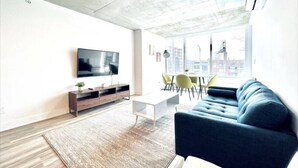 A modern living room with a flat-screen TV, a sofa, a coffee table, and a dining area with chairs. - Romema *201* - Condo Luxe 1 bdr (Montreal)