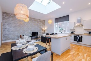 Une cuisine moderne dotée d’un îlot central, un coin repas avec une table dressée pour quatre personnes, et un espace salon avec un canapé et un téléviseur.