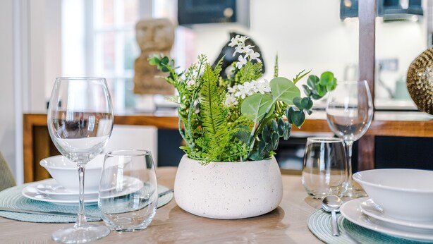 Una mesa de comedor dispuesta con cristalería transparente, un tazón blanco moteado y una maceta con follaje verde y flores blancas.