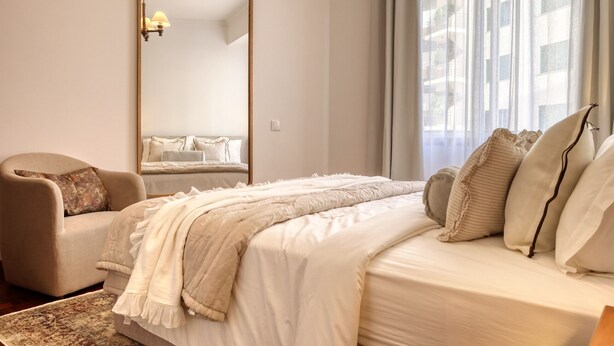 A bedroom with a large mirror, a bed with white bedding, a beige armchair, and a view of buildings through sheer curtains. - Elvira's Corner, a Home in Madeira (Funchal)