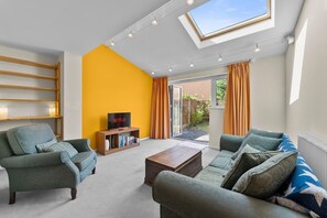 Una sala de estar con sofá, sillón y mesa de centro, un mueble para la televisión con libros, y un ventanal con cortinas.