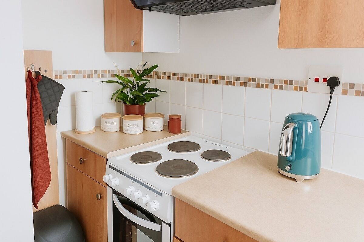 A compact kitchenette with a stove, oven, and a kettle. There are jars labeled 'SUGAR', 'COFFEE', and 'TEA' on the counter. A potted plant is on the counter, and a red towel hangs on the wall.