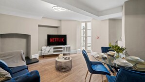 Een moderne woonkamer met een flatscreen televisie, een open haard, een eethoek met blauwe stoelen en een glazen tafel, en een groot raam dat natuurlijk licht binnenlaat.
