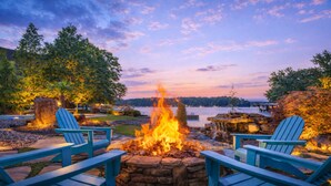 Ein Lagerfeuer, umringt von Adirondack-Stühlen, vor einer Kulisse aus Bäumen und einem See bei Dämmerung.