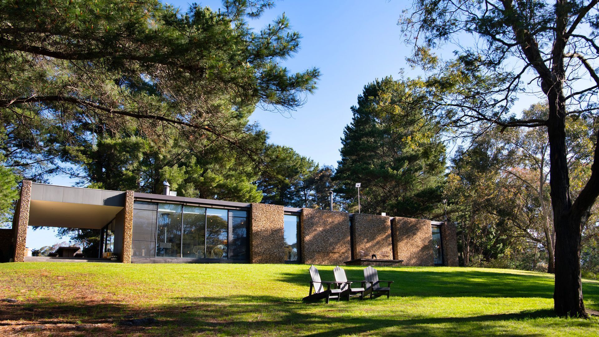 Un bâtiment moderne doté de grandes fenêtres en verre et d’une façade en pierre, entouré d’une pelouse et d’arbres.