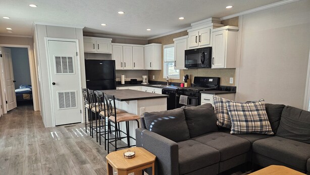 A modern kitchen with white cabinets, a black refrigerator, and a microwave. There is a dining area with a table and chairs. A gray sofa and a small wooden table are in the living room. - Lakefront retreat on West Battle Lake cozy, modern, and steps from the water! (Battle Lake)
