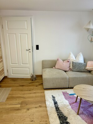 Ein Zimmer mit Holzboden, einem beigefarbenen Sofa mit Kissen, einer weißen Tür und einem kleinen Tisch mit runder Platte.