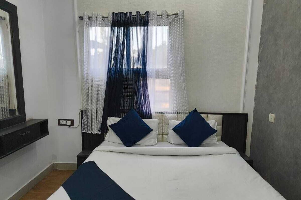 Un dormitorio con una cama, dos almohadas, una mesita de noche, un espejo y una ventana con cortinas.