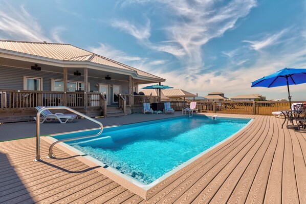A swimming pool with a wooden deck, lounge chairs, and umbrellas. - Luxurious Coastal Home with Heated Pool and Views (Dauphin Island)