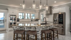 A modern kitchen with white cabinets, a large island with bar stools, stainless steel appliances, and pendant lighting. - Luxurious Coastal Home with Heated Pool and Views (Dauphin Island)