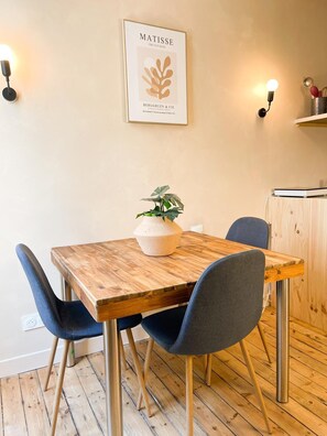 A dining area with a wooden table, two chairs, a potted plant, and a framed artwork on the wall.