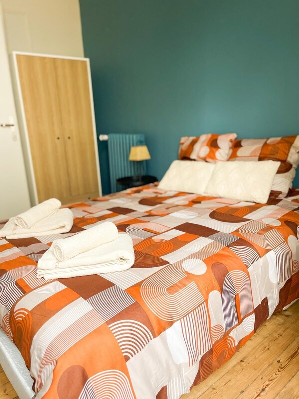 A neatly made bed with a patterned bedspread, two pillows, and folded towels on a wooden floor.