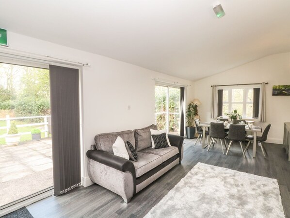 A modern living room with a grey sofa, a dining table with chairs, and a large window providing a view of the outdoors.