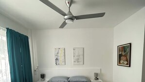 Une chambre à coucher comprenant un lit, un ventilateur de plafond, une table de chevet et une commode, ainsi qu’une fenêtre avec des rideaux.
