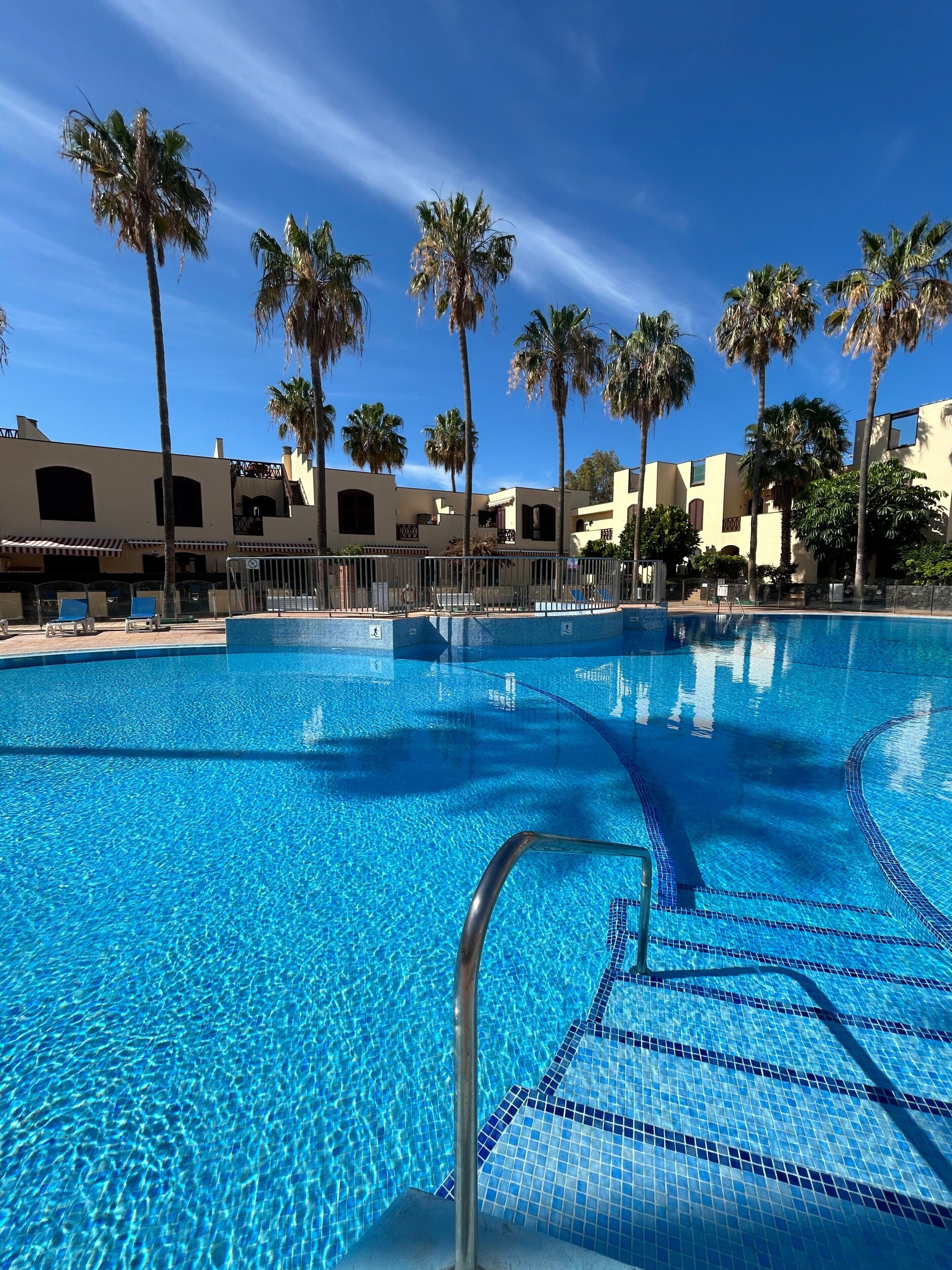 A clear blue swimming pool with a tiled edge, surrounded by palm trees and buildings with a Mediterranean architectural style.