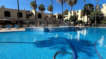 A clear blue swimming pool with a tiled edge, surrounded by palm trees and buildings with a Mediterranean architectural style.