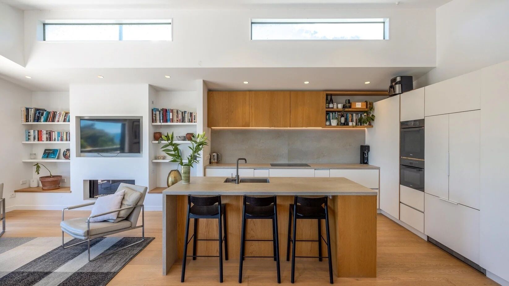 A modern kitchen with a central island, bar stools, a built-in oven, and a TV above the island.
