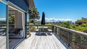 A wooden deck with a glass door leading to a balcony, a table with chairs, and a black umbrella. - Onetangi Seaview Residence (Waiheke Island)