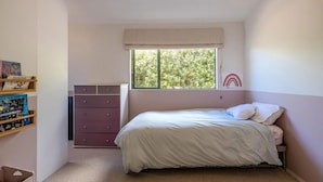 Ein Schlafzimmer mit Bett, Kommode, Fenster mit Blick auf Bäume und einem Bücherregal.